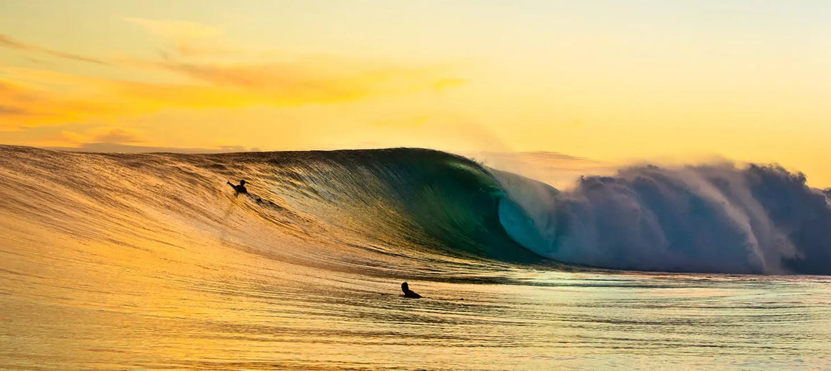 Surfers riding a large wave at sunset with a colorful sky.