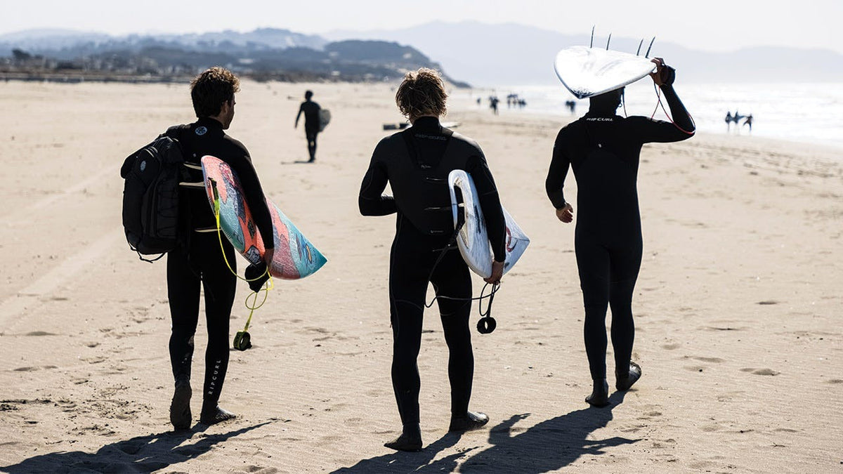 3 people walking along the beach wearing Rip Curl wetsuits
