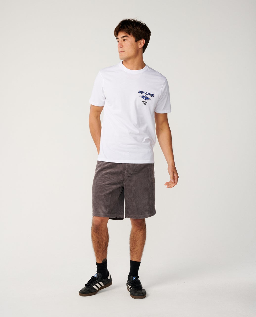 A young man stands in a white t-shirt and gray corduroy shorts, wearing black sneakers and socks, looking to the side.