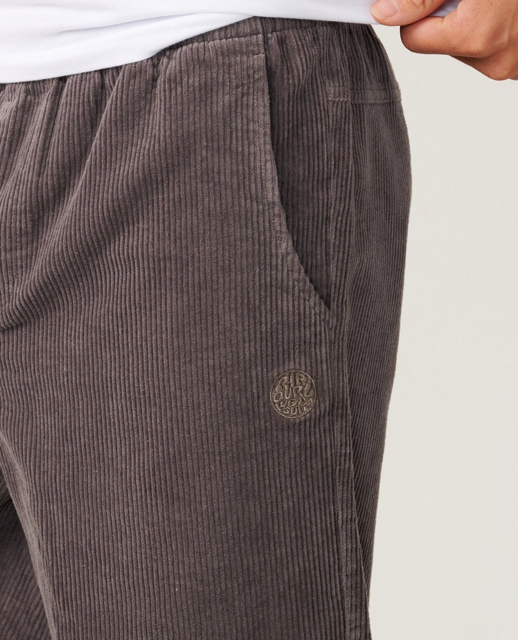 Close-up of textured, dark brown corduroy pants with a side pocket and a circular logo on the front.