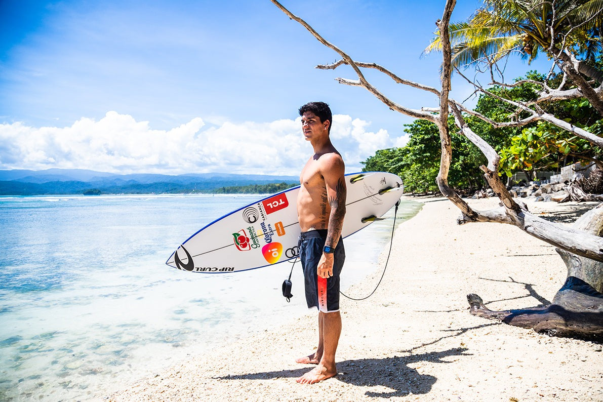 Man holding a surfboard on a tropical beach with palm trees and clear blue water.