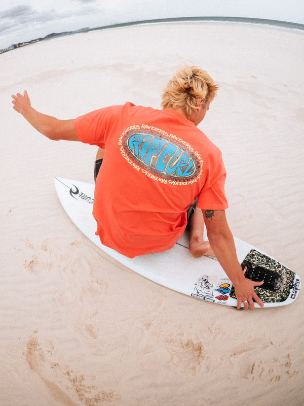 Person sitting on a surfboard with a colorful logo on an orange shirt, standing on a sandy beach.