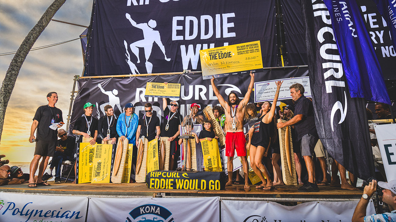 Group of people on a stage with surfboards, celebrating an event with Eddie Would Go and Rip Curl banners.