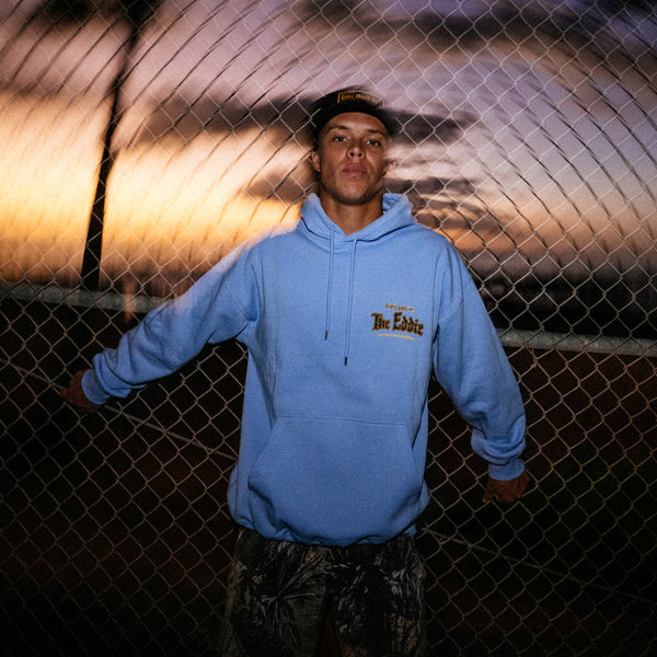 Person wearing a blue hoodie with a logo in front of a chain-link fence.