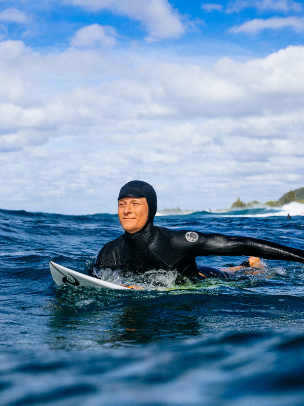 A man in a black full-body wetsuit floating on a surfboard in the ocean under a blue sky with white clouds.
