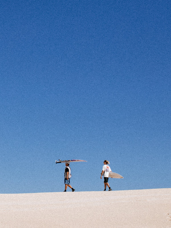 Two people walking on a beach with surfboards under a clear blue sky.