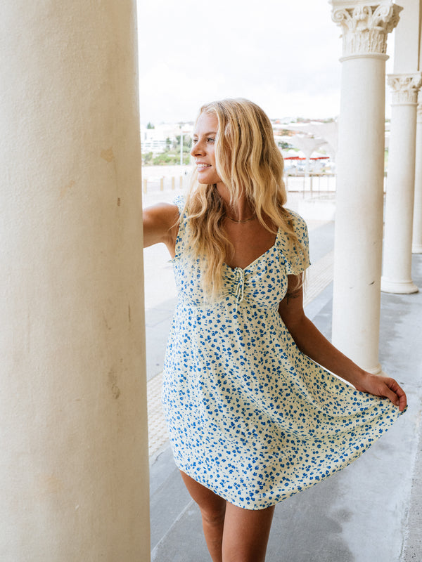 Woman in a floral dress standing in a sunlit outdoor setting with columns.