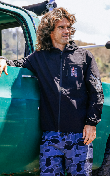 Person wearing a black jacket with a logo and blue patterned pants standing next to a green vehicle.