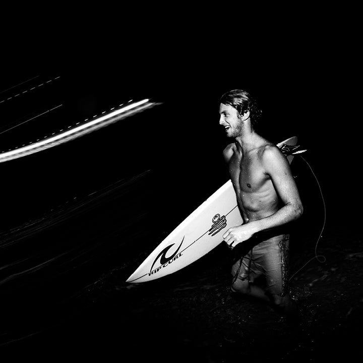 Man holding a surfboard at night with a car in the background