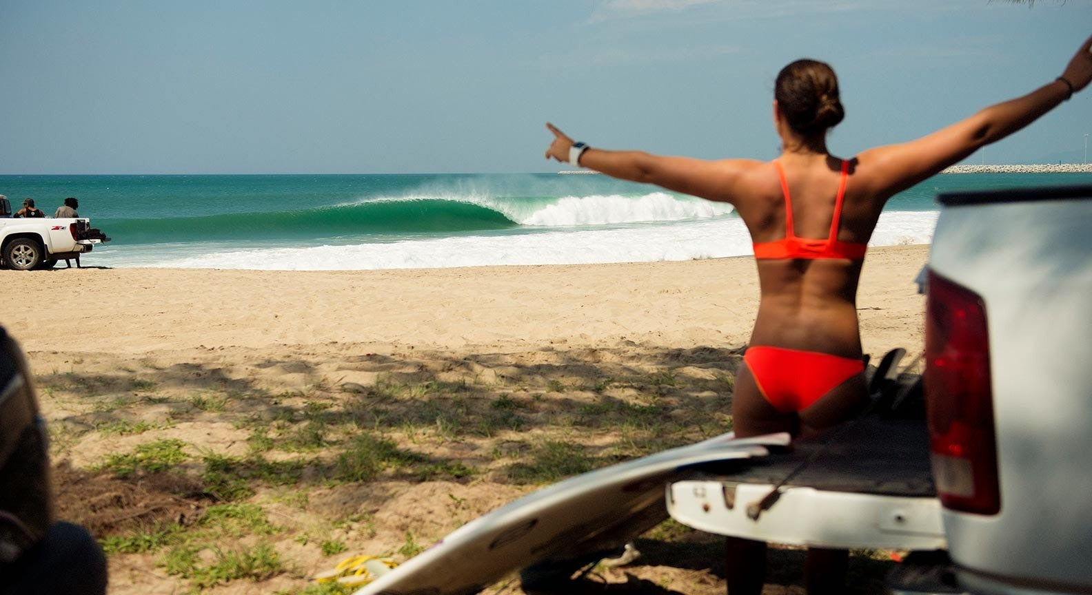 Woman in red bikini sitting on a car with arms outstretched on a beach, looking at the ocean.