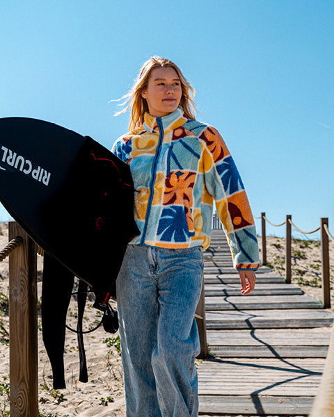 Person wearing a colorful jacket and jeans holding a surfboard on a wooden path with a clear blue sky.