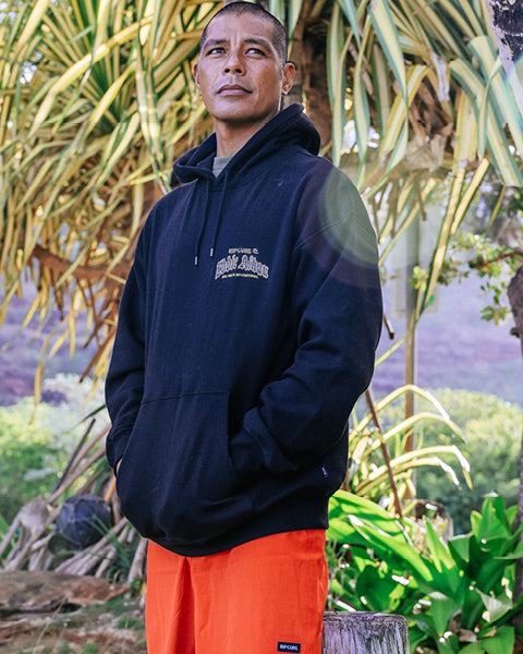 Man wearing a navy hoodie with a logo and orange pants standing in a natural setting with plants and trees.