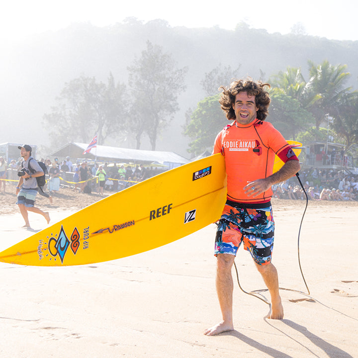 Man holding a yellow surfboard on a beach with trees and people in the background