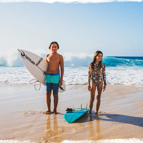 Two people standing on a beach with a surfboard and a kayak, ocean waves in the background.