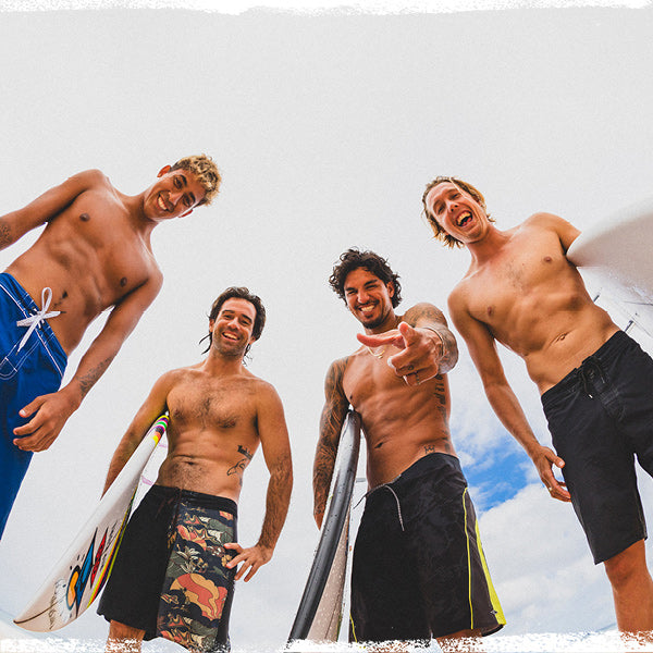 Four shirtless men standing on a beach with surfboards, smiling and posing for the camera.