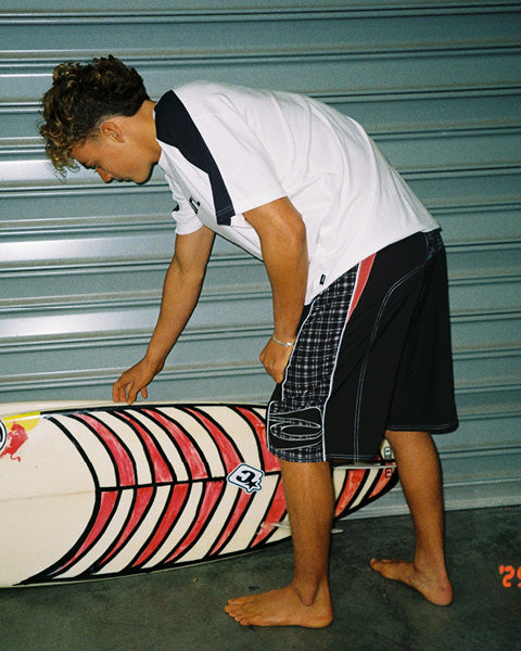 A man in a white and navy t-shirt and black plaid shorts bends over next to a surfboard with red, black, and white stripes, set against a gray metal garage door background.