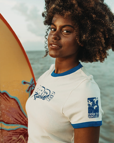 A woman with curly hair wearing a white short-sleeve ringer t-shirt with blue trim stands by the ocean holding a surfboard. The shirt features a blue graphic on the chest and a 'Farm to Front' sleeve logo.