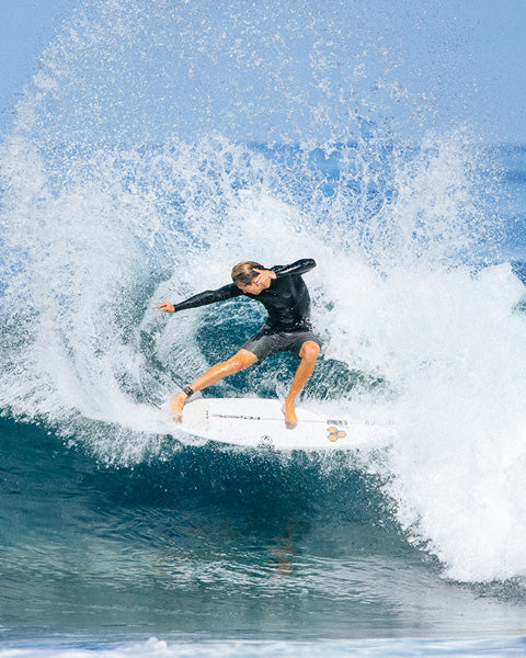 A surfer in a black wetsuit rides a white surfboard on a breaking ocean wave, with large white spray and blue water filling the frame.