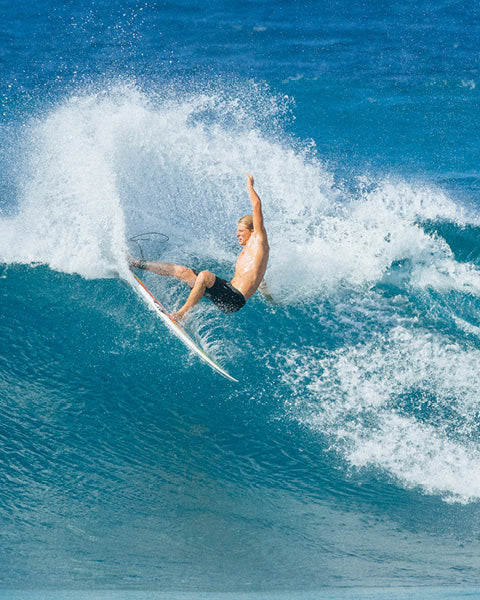 A shirtless male surfer riding a large blue ocean wave with white spray, wearing black swim trunks and balancing on a white surfboard.