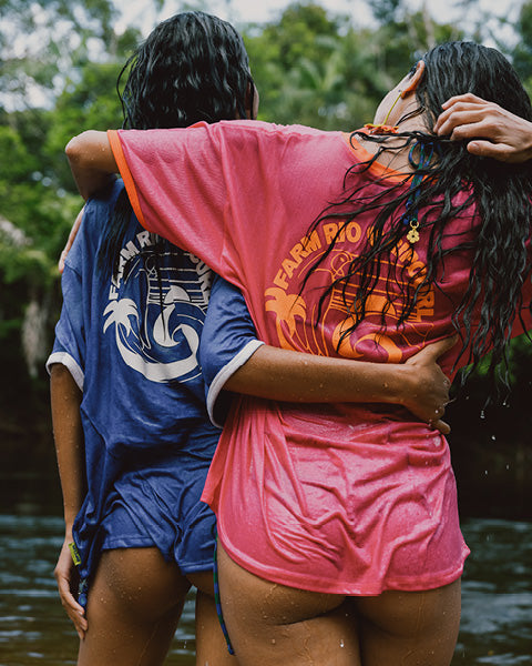 Two people embracing outdoors near water, wearing oversized matching swim shirts and shorts. One shirt is bright pink with orange graphic text and the other is blue with a white anchor graphic.