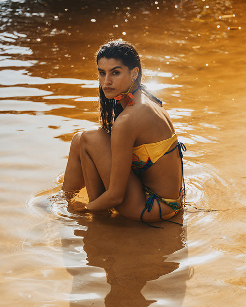 A woman sitting in shallow water at the beach, wearing a yellow bikini with multicolor side-tie details. The water is golden and reflective under warm sunlight.