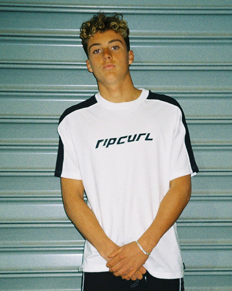 A young man with curly blonde hair wearing a white Rip Curl t-shirt with black shoulder panels and the brand name printed across the chest, standing in front of a gray metal shutter.