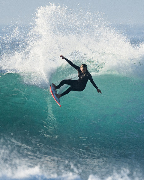 A surfer in a black wetsuit rides inside a large turquoise ocean wave with white spray, captured mid-action against a bright sky.