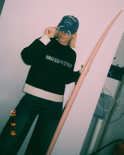 A person wearing a black Rip Curl sweater with white trim, dark pants, and a blue patterned cap, holding a long pink surfboard against a plain wall.