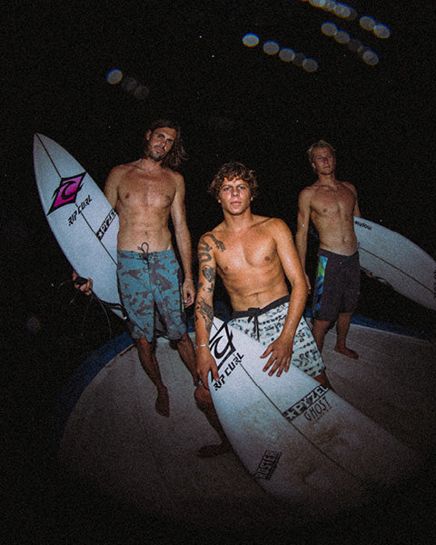 Three shirtless men standing on a deck at night, each holding a surfboard. They are wearing swim trunks, and the surfboards have visible logos including Swamfins and Rip Curl.