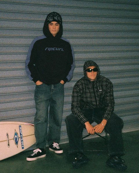 Two men posing in front of a gray metal garage door. The standing man wears a black and gray Rip Curl hoodie, blue jeans, and black-and-white sneakers, with a surfboard visible at his side.