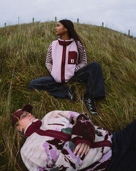 Two models wearing matching knit outerwear sets are posed on a grassy hill under an overcast sky. One model sits cross-legged while the other lies down, showcasing the soft textures and patterns of the garments.