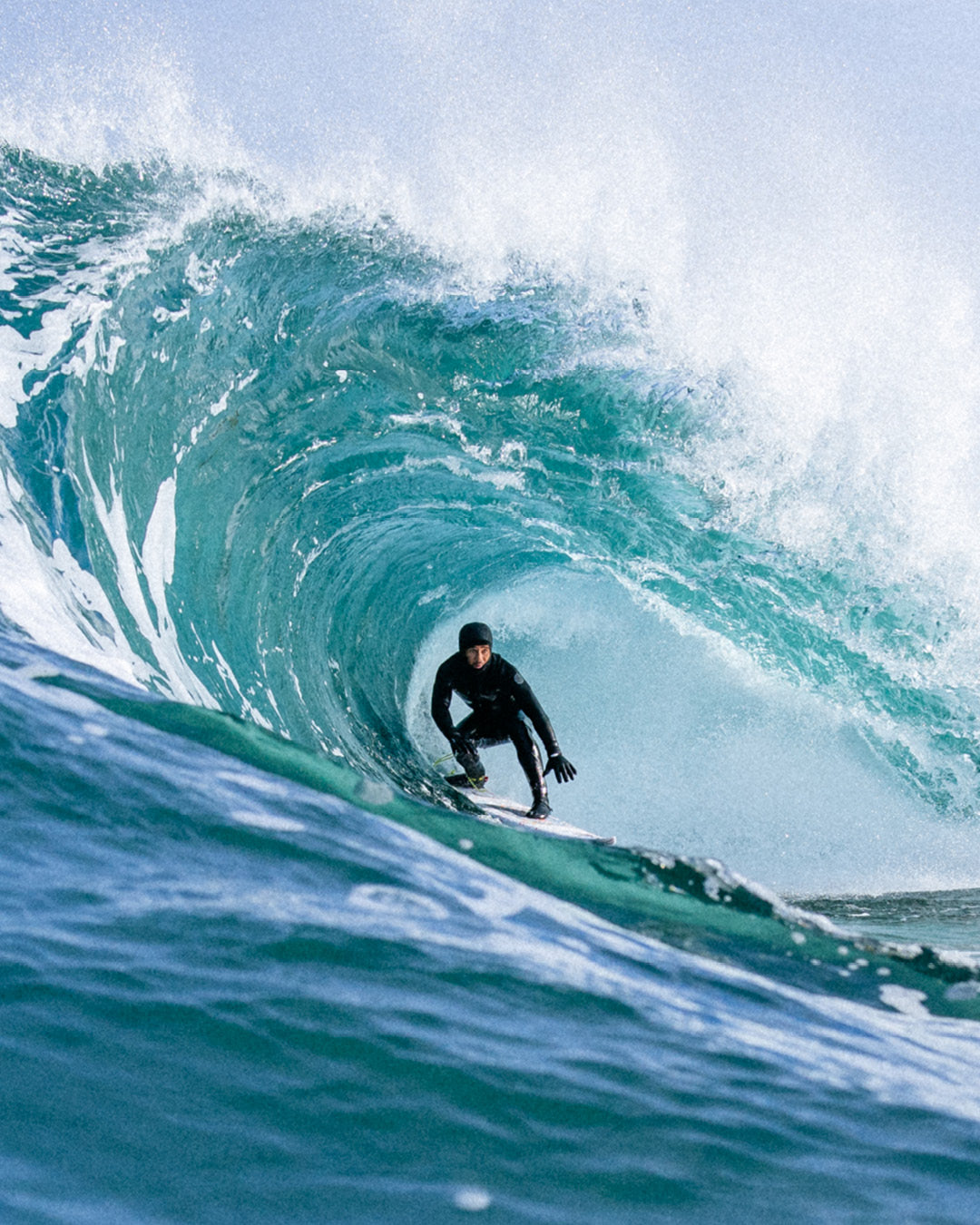 A surfer in a black wetsuit rides inside the barrel of a large turquoise ocean wave with white spray, photographed from a low angle.