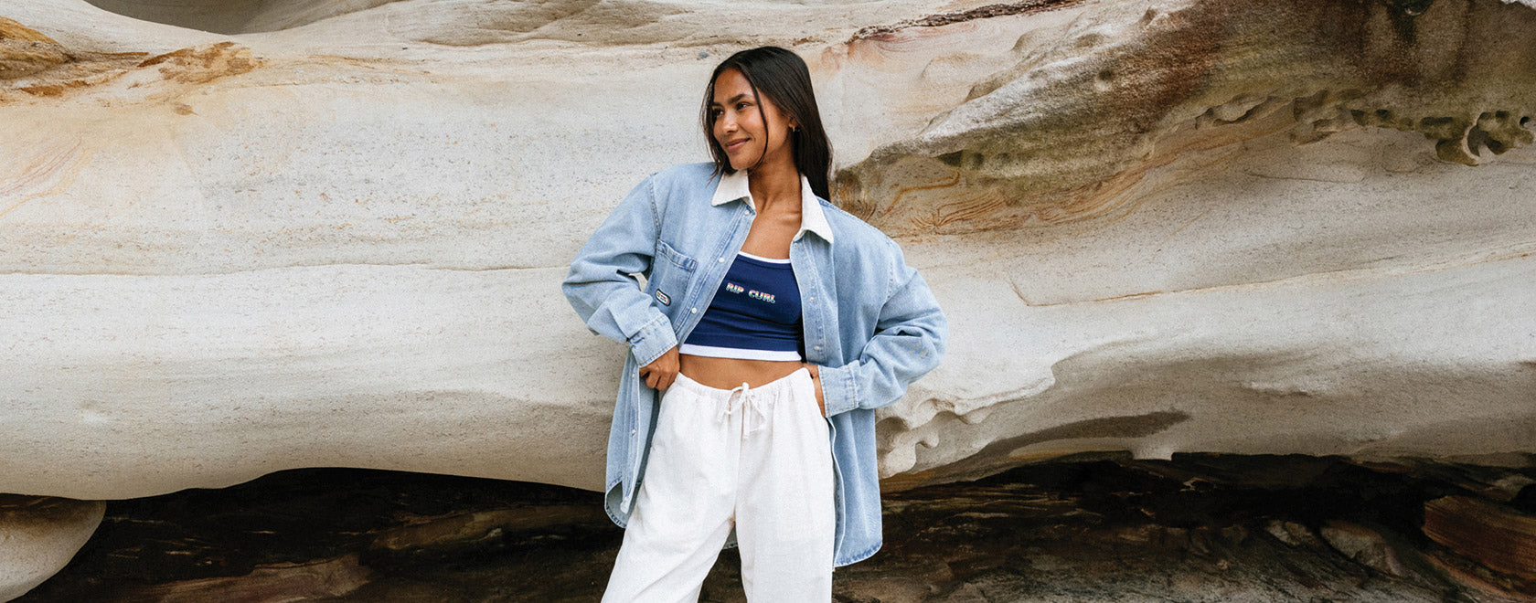 Woman standing in front of a large rock formation wearing a light blue denim jacket and white pants.