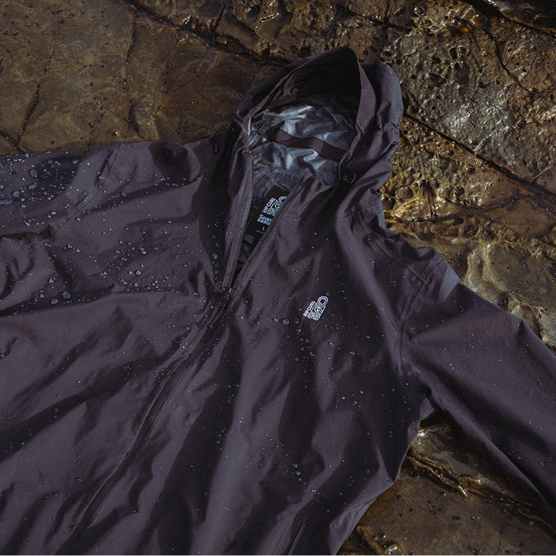 A dark waterproof hooded jacket with water droplets on the surface, laid flat on a wet rocky surface. The jacket has a small white logo on the left chest.
