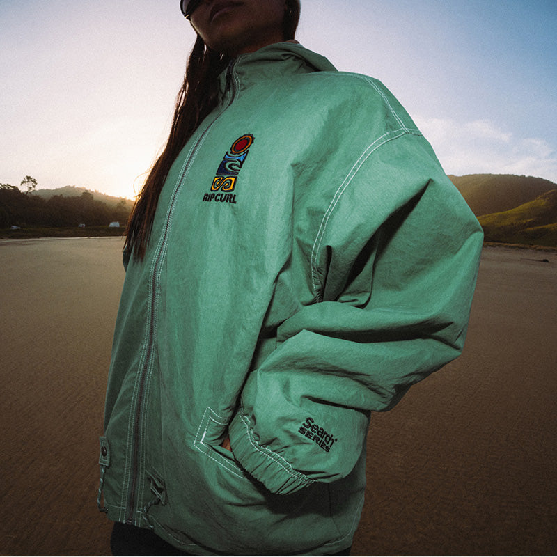 A person wearing an oversized mint green Rip Curl jacket in an outdoor beach setting at sunset. The jacket features a colorful Rip Curl logo on the left chest and 'SCORPION' printed on the right sleeve cuff.