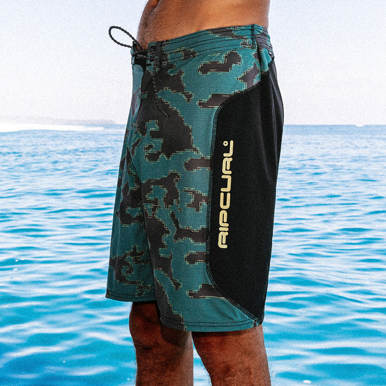 A close-up side view of a person wearing Rip Curl board shorts with a teal and black camouflage pattern and a solid black side panel, standing against a blue ocean background.