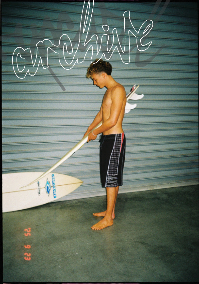 A shirtless man standing barefoot in front of a gray metal garage door, holding a white surfboard and paddle. The surfboard has blue branding and the man is wearing dark board shorts with a red stripe.