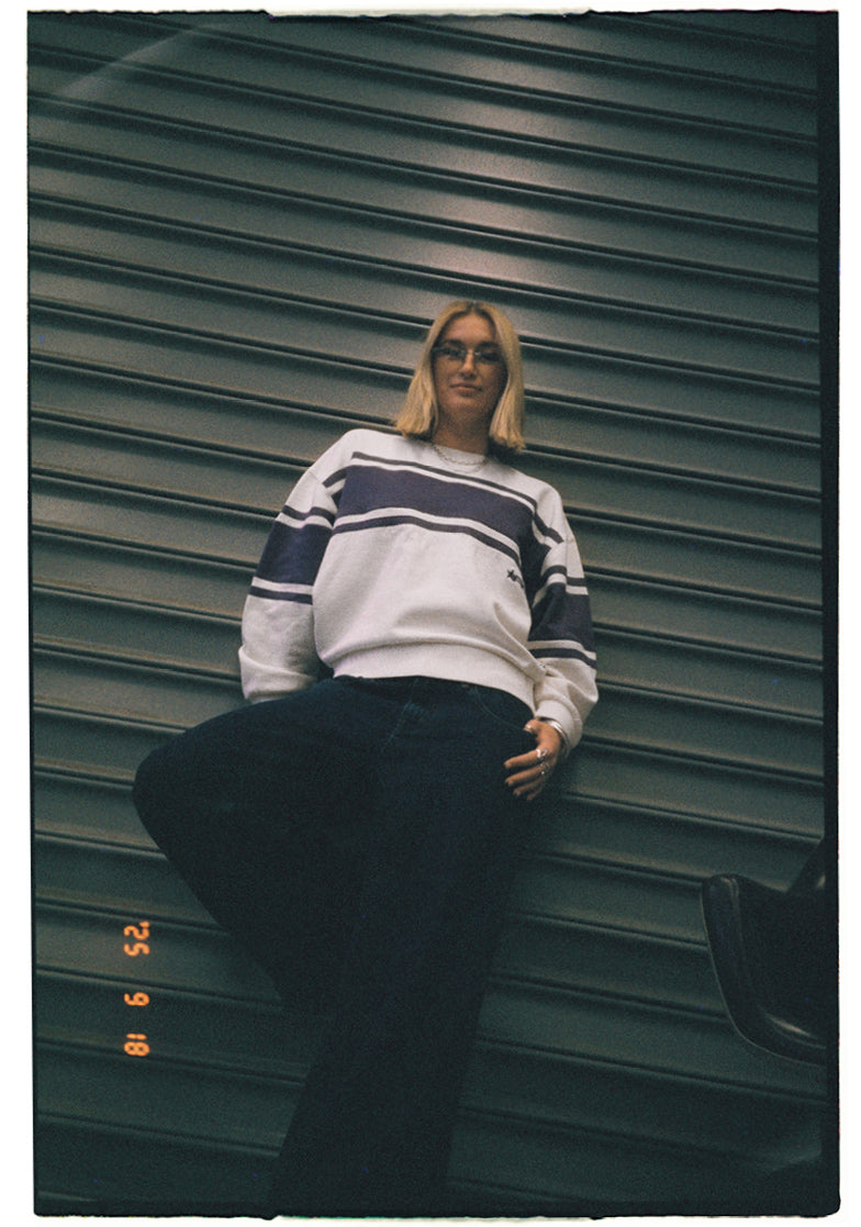 Person wearing a striped sweater and dark pants against a green corrugated metal wall.