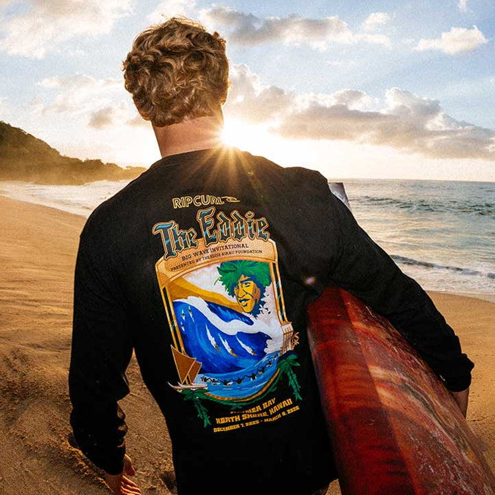 Person holding a surfboard on a beach with 'The Eddie' logo on their shirt