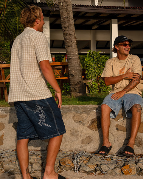Two men are having a conversation outdoors with a palm tree and wooden furniture in the background.