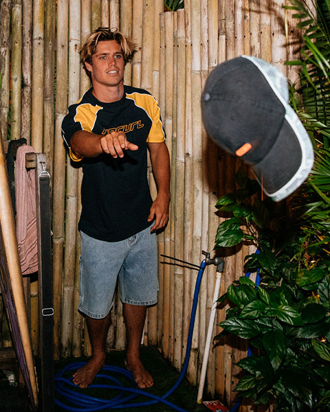 Man standing next to a bamboo wall with a cap and hose, surrounded by plants.