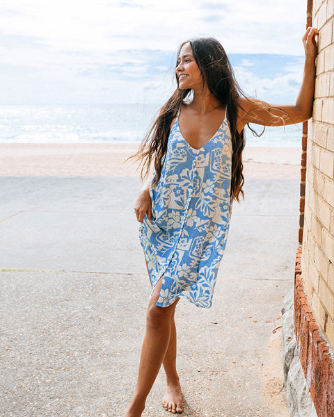 Woman in a blue floral dress standing by a brick wall with a beach in the background