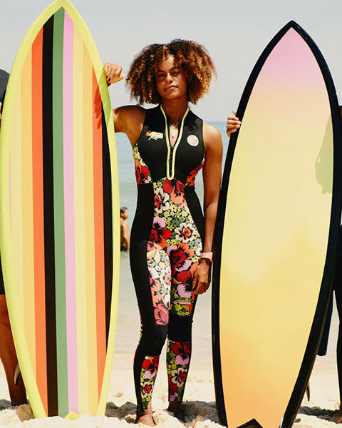 Woman in a floral wetsuit holding two surfboards on a beach