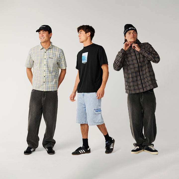 Three male models standing side-by-side in a studio setting, showcasing different streetwear outfits. The outfits include a light green and white plaid short-sleeve button-up shirt, a black t-shirt with a blue graphic, and a dark brown and black plaid button-up shirt, paired with dark gray jeans, light blue denim shorts, and black sneakers.