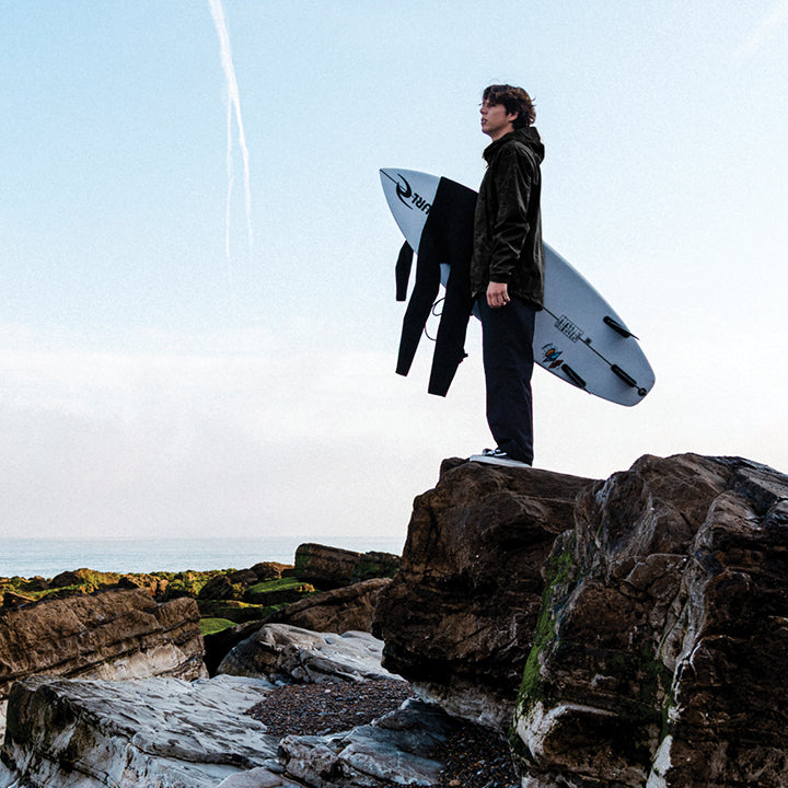 A surfer stands on rocky coastal ground holding a white surfboard and a black wetsuit under their arm, with the ocean and pale sky in the background.