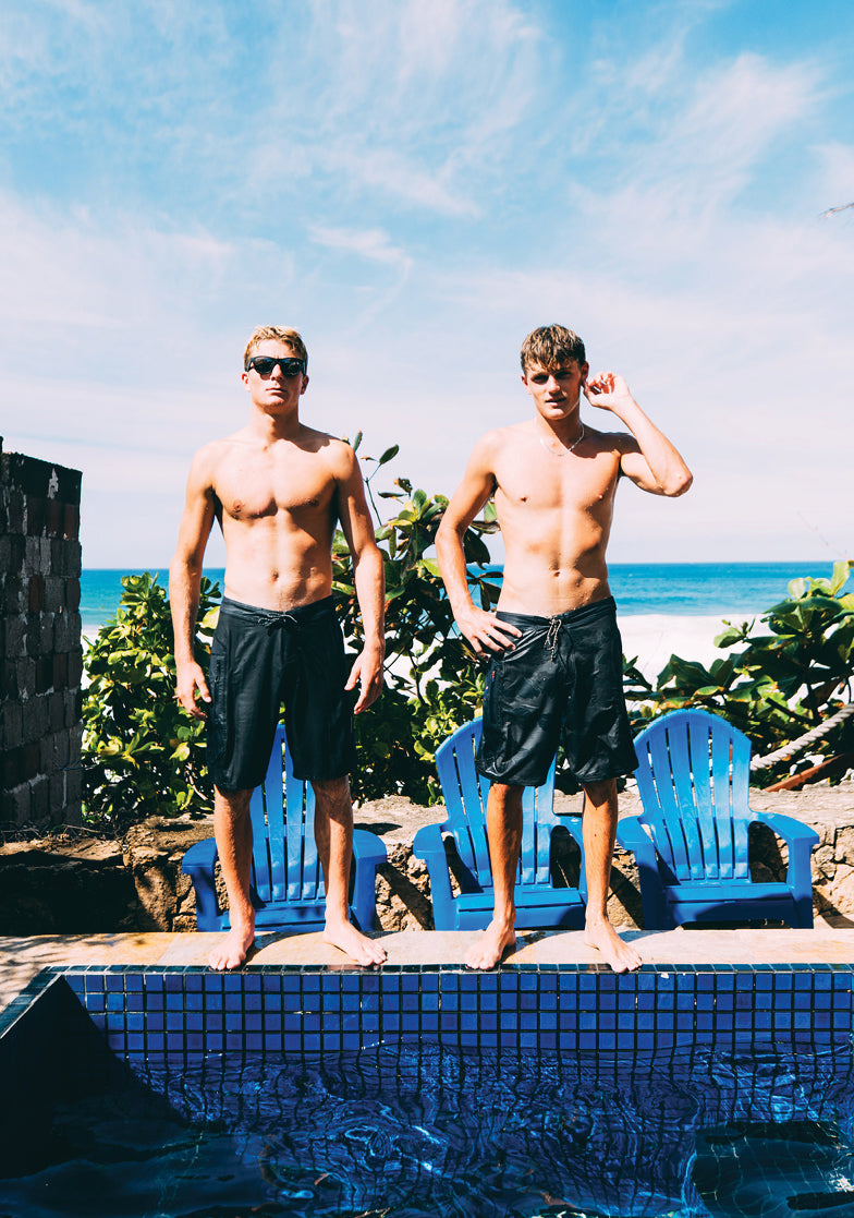 Two shirtless men standing by a pool with a beach and ocean view.
