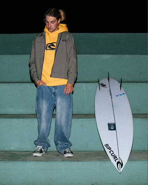 Person standing next to a Rip Curl surfboard on a green wall.