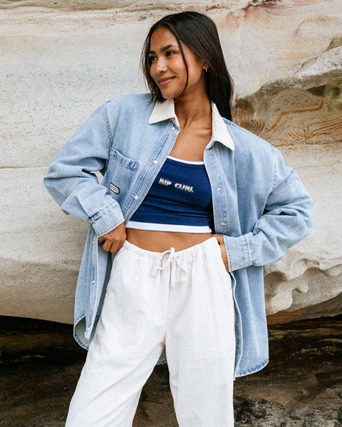 Woman wearing a blue denim jacket, blue crop top, and white pants against a stone wall.