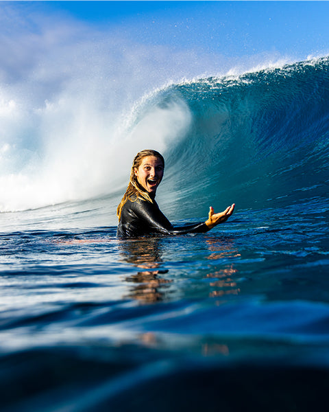Person in a wetsuit paddling in the ocean with a large wave in the background