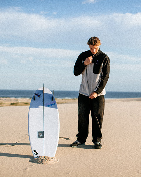 Person standing on a beach holding a surfboard with a clear sky and ocean in the background